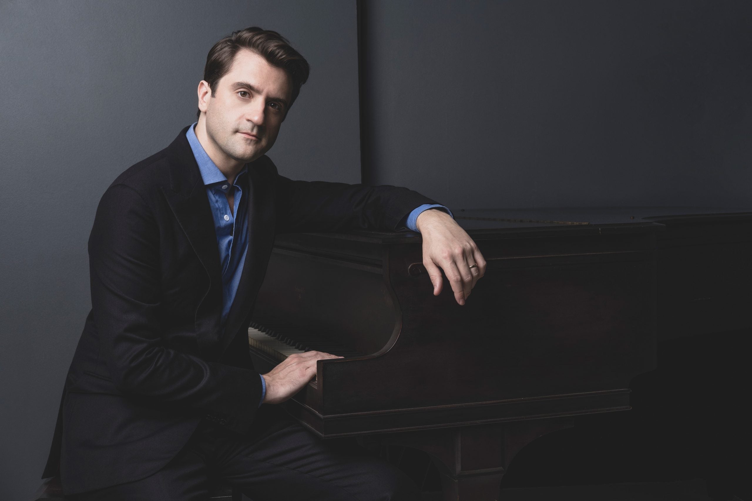 A man, wearing a black suit and blue oxford shirt, sitting at a piano with a grey background.