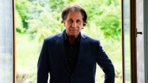 A man with mid-shoulder length hair, wearing a black suit, standing in front of foliage