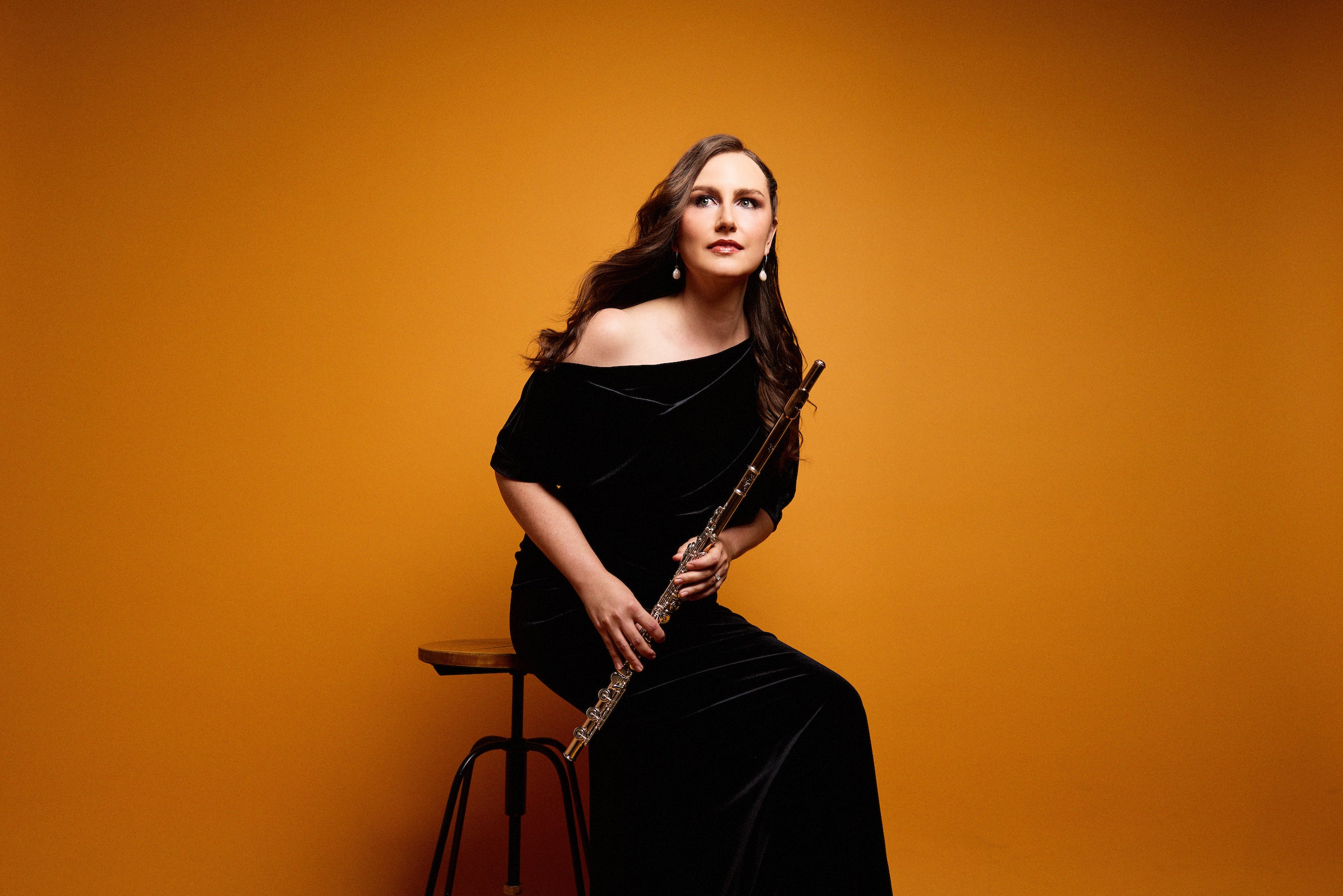 Woman in black dress holding a flute seated on a stool in front of an orange background.