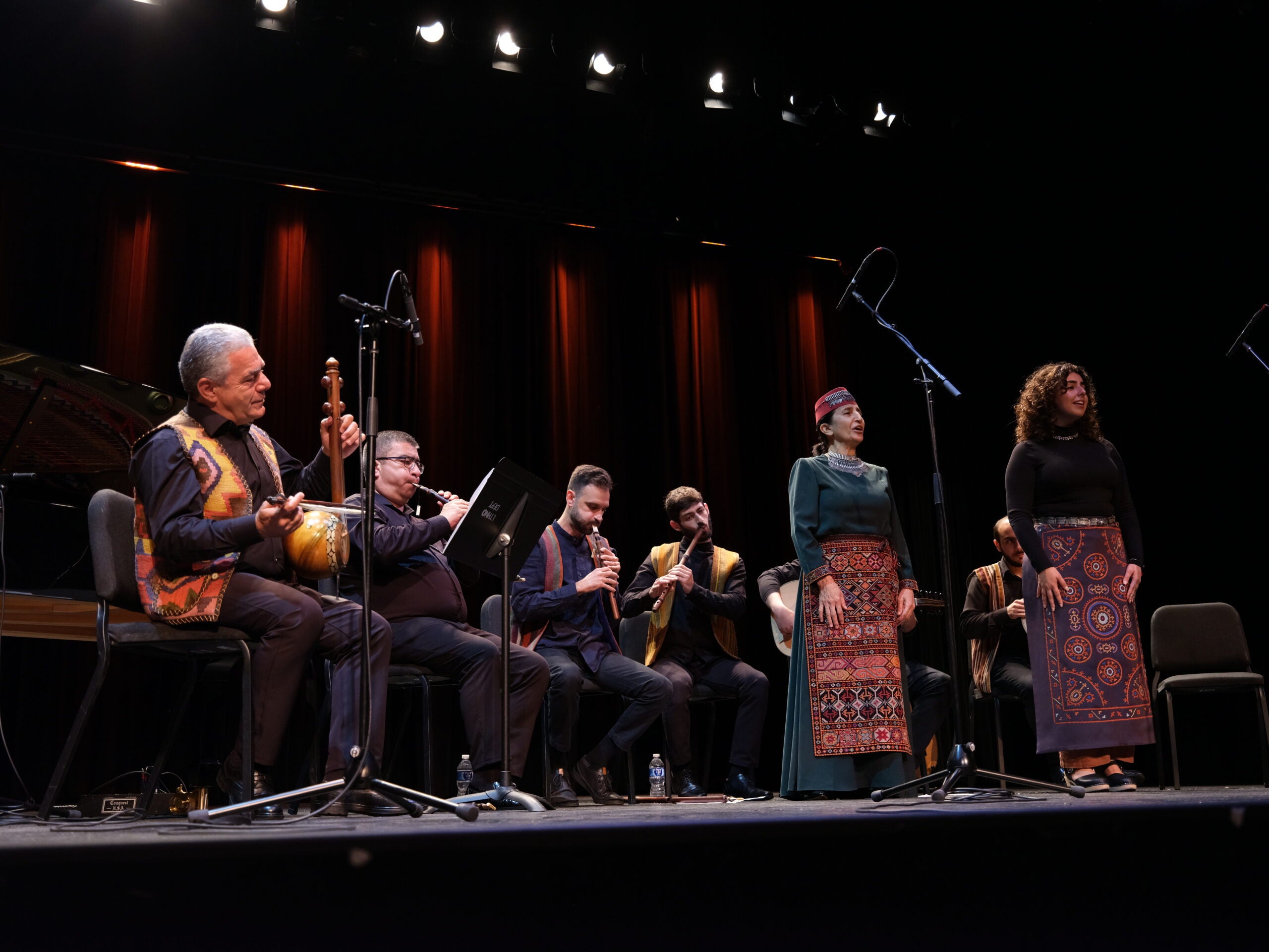 A group of Armenian folk ensemble musicians performing on stage, dressed in vibrant traditional costumes featuring patterned vests and aprons. The musicians are seated in a half circle, with various traditional instruments such as the kamancha, duduk, blul, oud, and dhol being played. In the center, there are two vocalists singing.