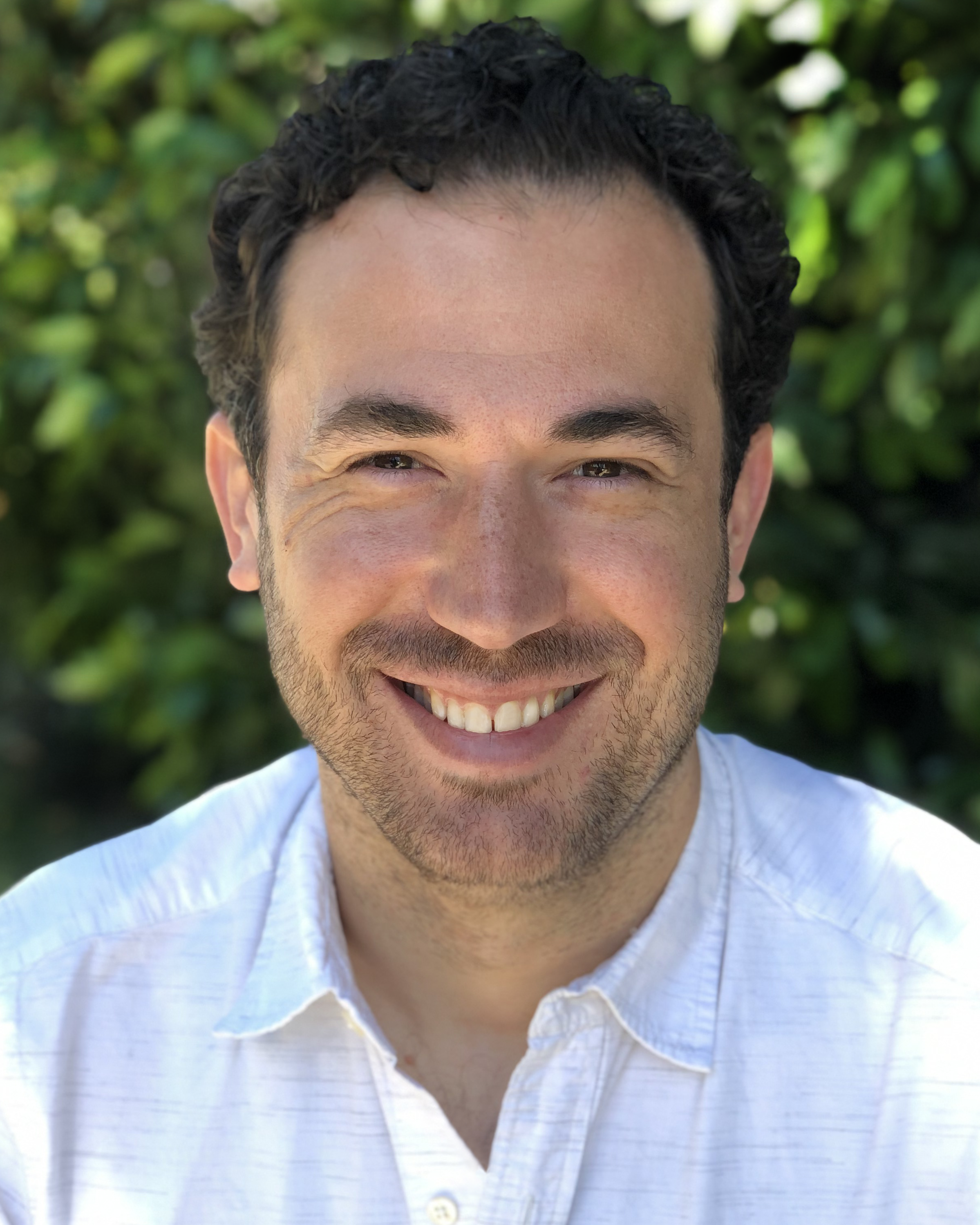 man with dark curly hair, smiling and wearing a white shirt