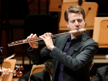 Man in black suit, with short hair, playing flute.