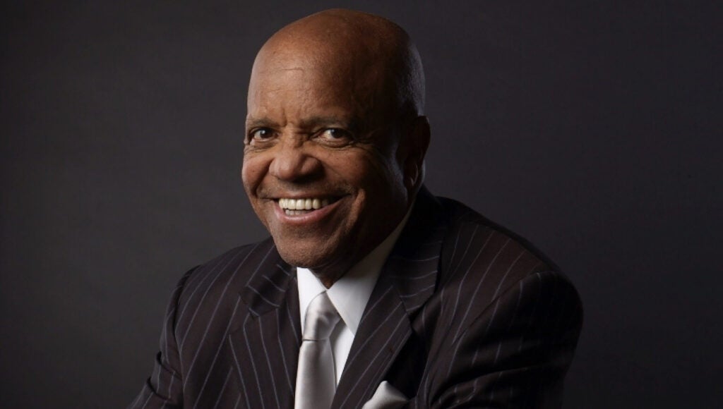 Berry Gordy, African American man sitting with hands clasped in front of him, dressed in a dark suit with a white shirt and gray tie.