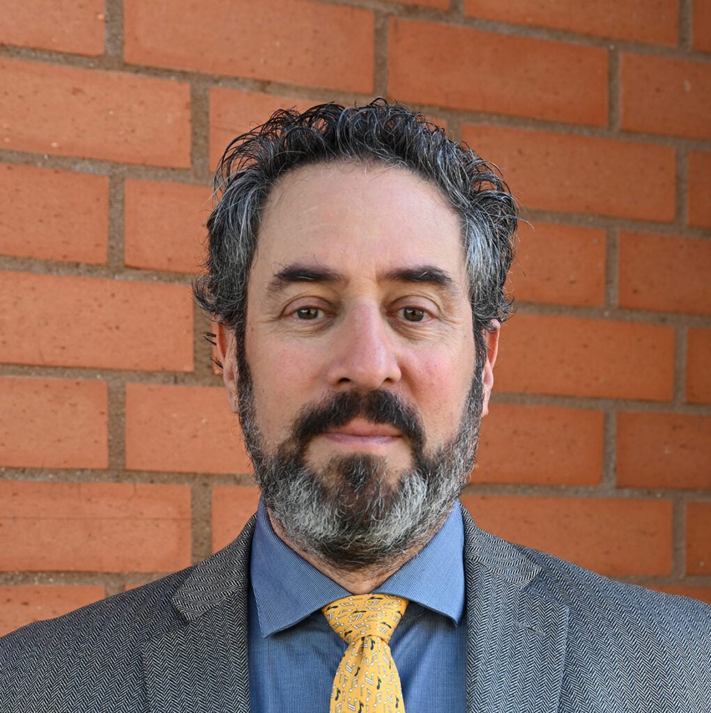 man in a gray sportscoat with yellow tie standing against a brick wall.