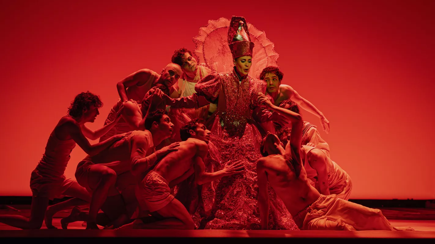 An operatic scene that involves a number of people converging on a character wearing a pope's hat, on a red background.