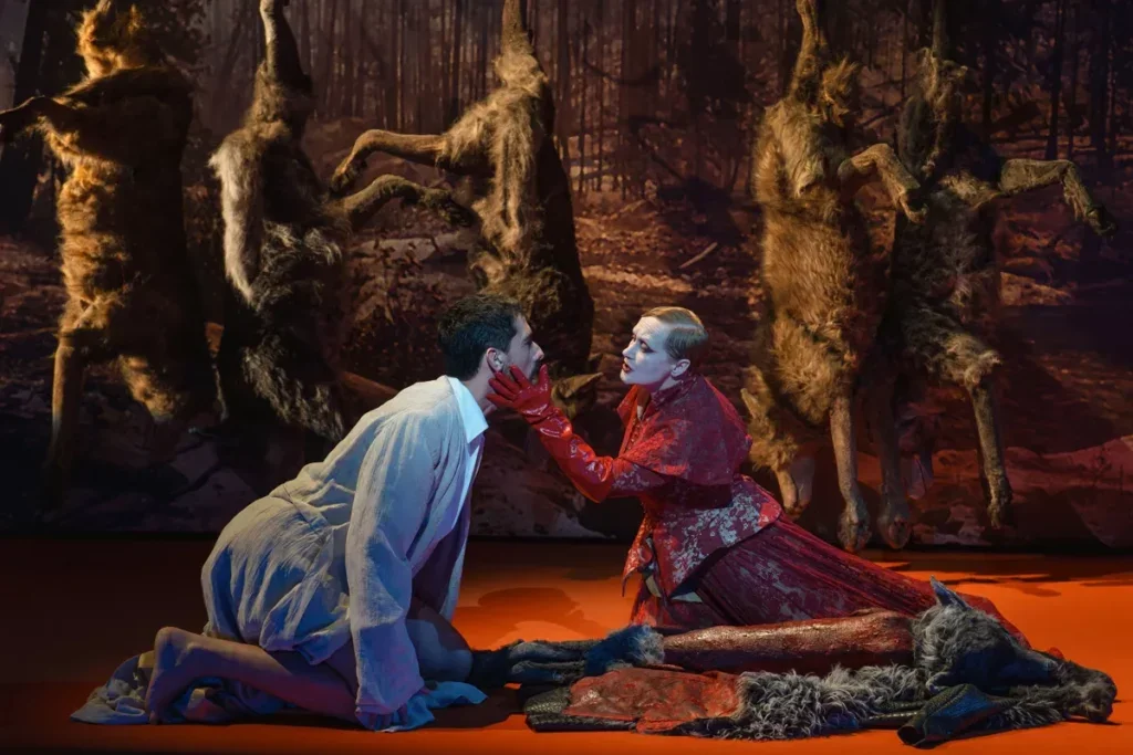 two characters kneeling, one with her hand on the face of a man, beneath hanging wolf pelts.