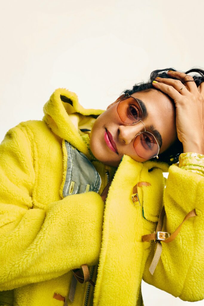 Woman in yellow sweatshirt and sunglasses with her head tilted to her left side.