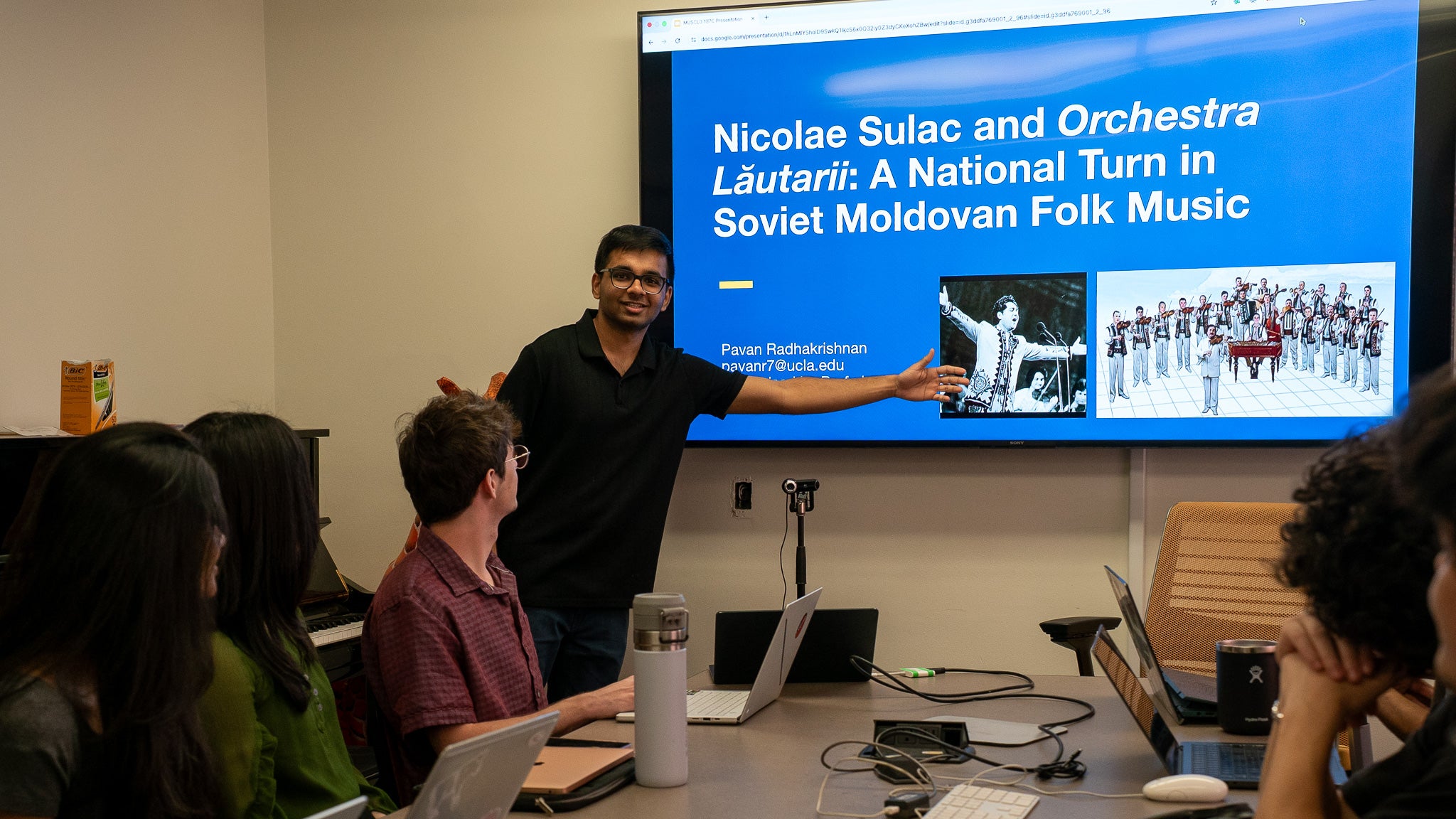 Man in short sleeved black t-shirt pointing to a powerpoint.