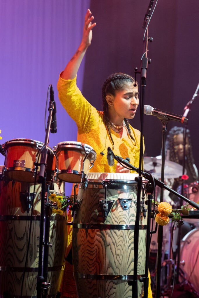 Woman in yellow shirt sitting at a drum set with a microphone.