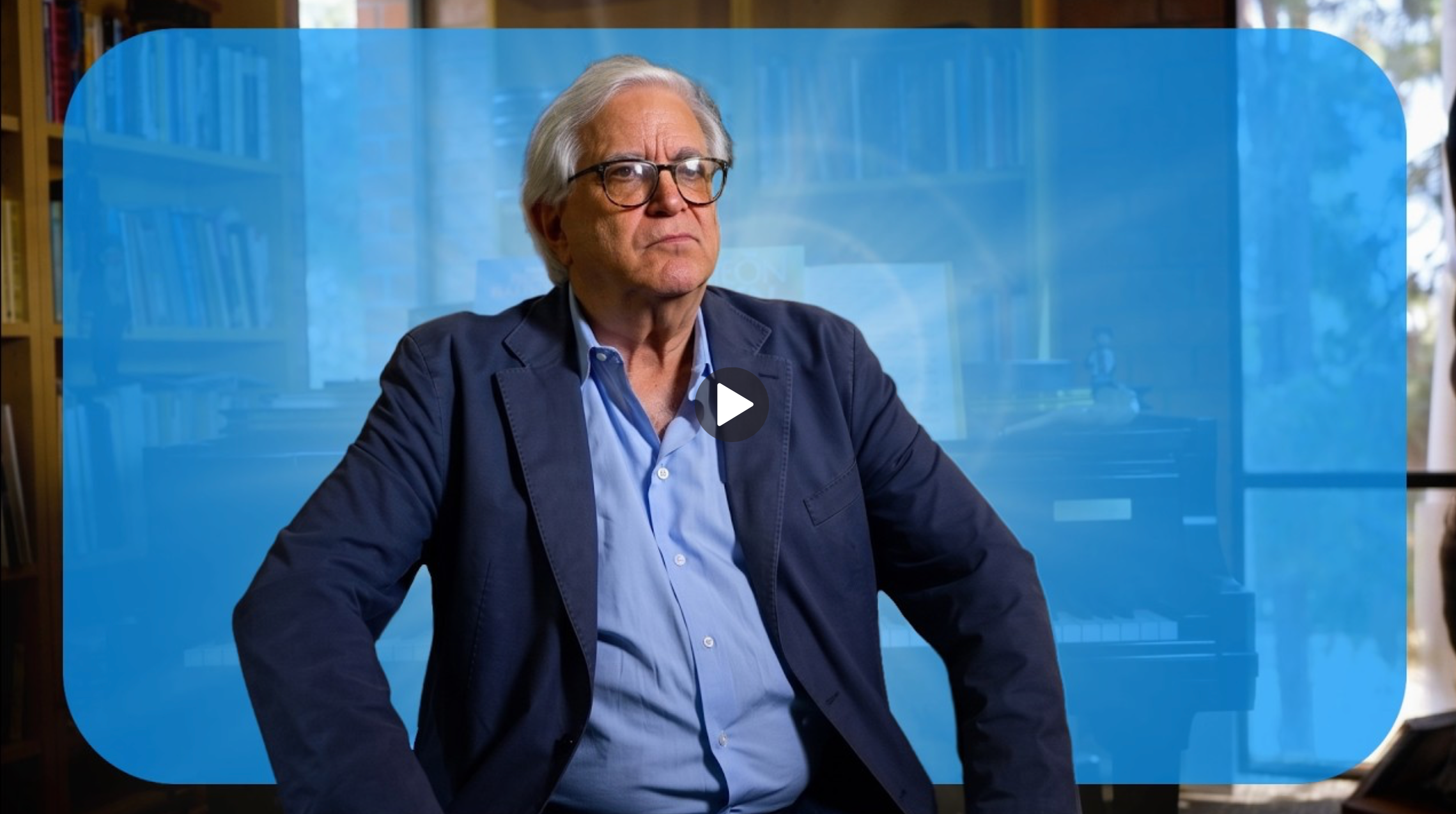 Man with white hair and glasses in a blue blazer and blue shirt against a blue UCLA background.