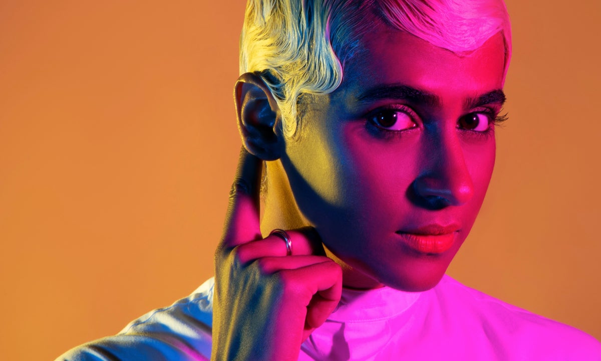 Woman with short blonde hair standing with finger pointing to her ear.