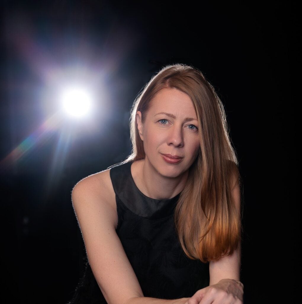 Woman in black dress standing against a black background with a stage light shining toward the camera.