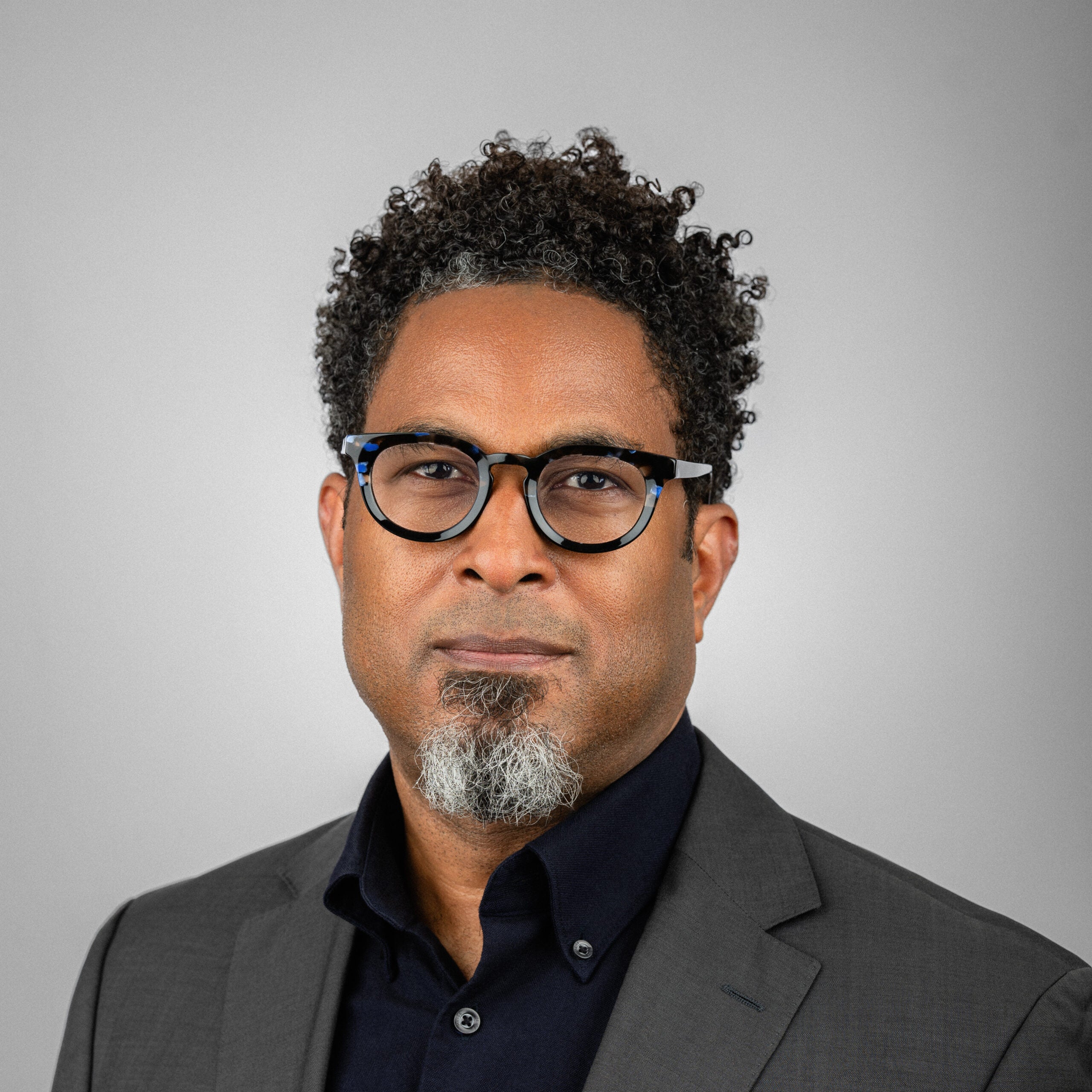 Man with black hair in Black glasses and a gray coat in front of a gray background.