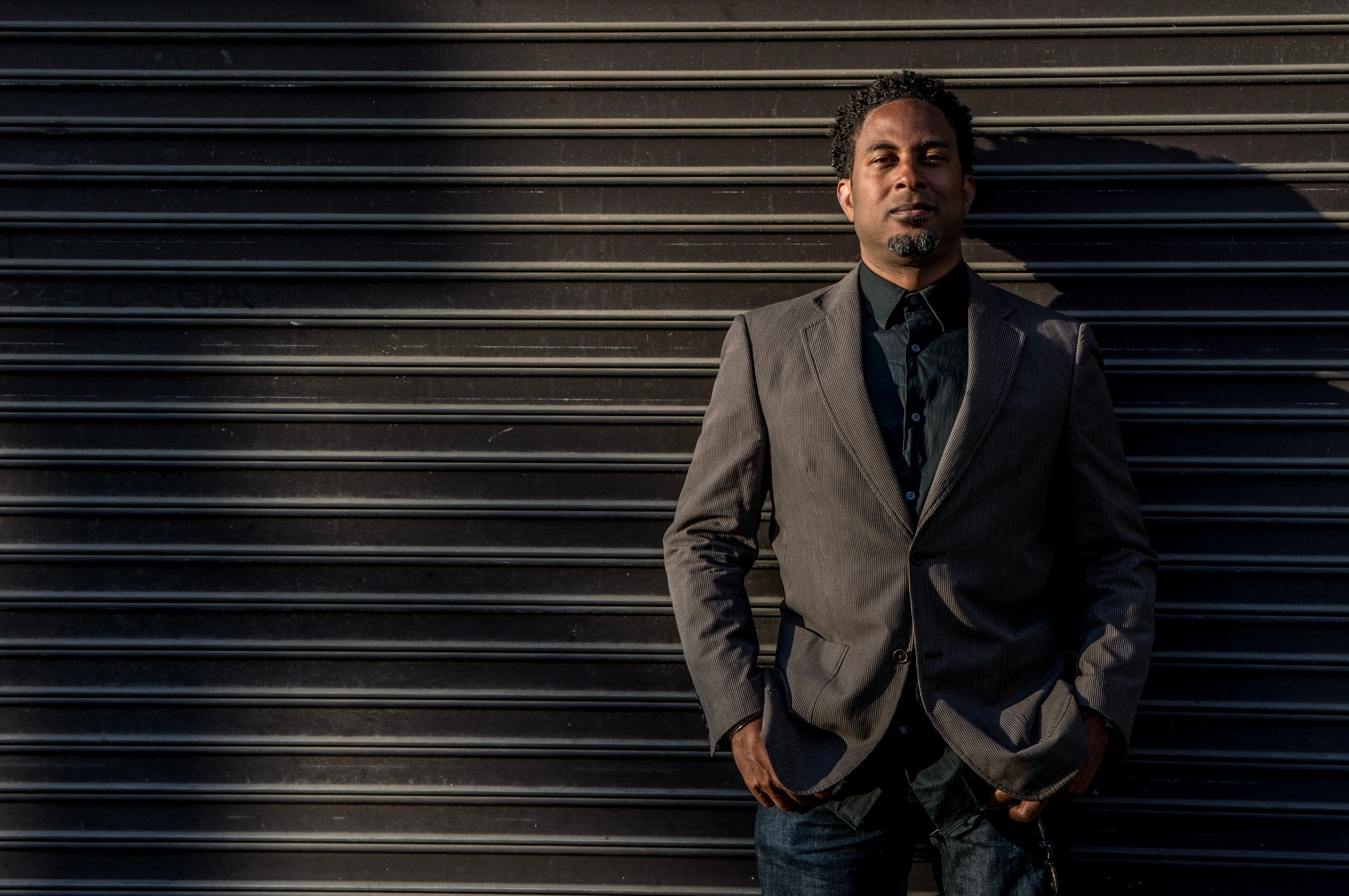 Man in gray suit jacket and black shirt standing against a wall.