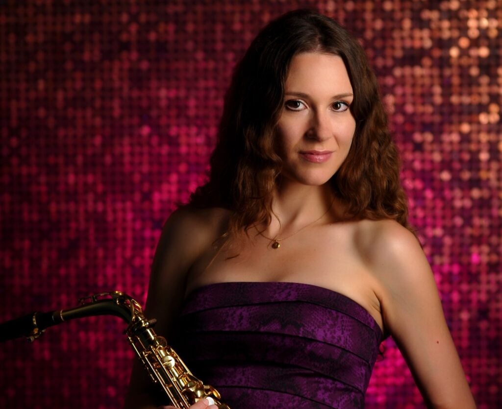 woman in red strapless dress holding an alto saxophone standing against a red background.