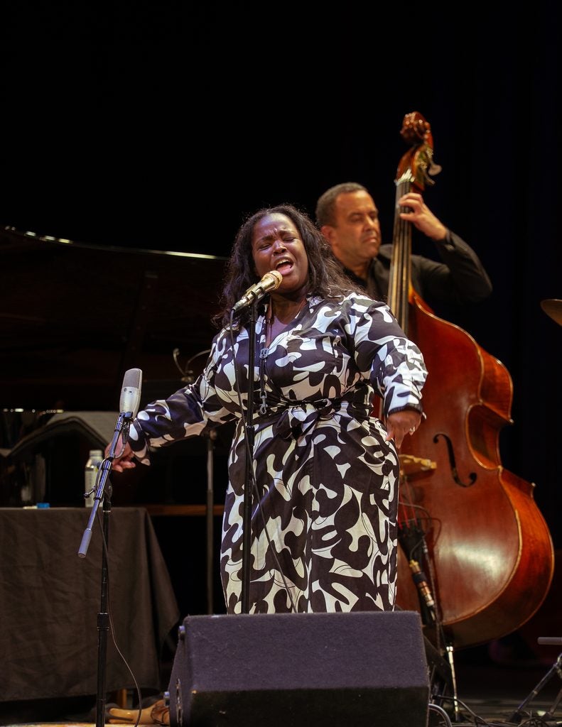 Woman in a black and white print dress singing at a microphone, a man on stand up bass is playing behind her.