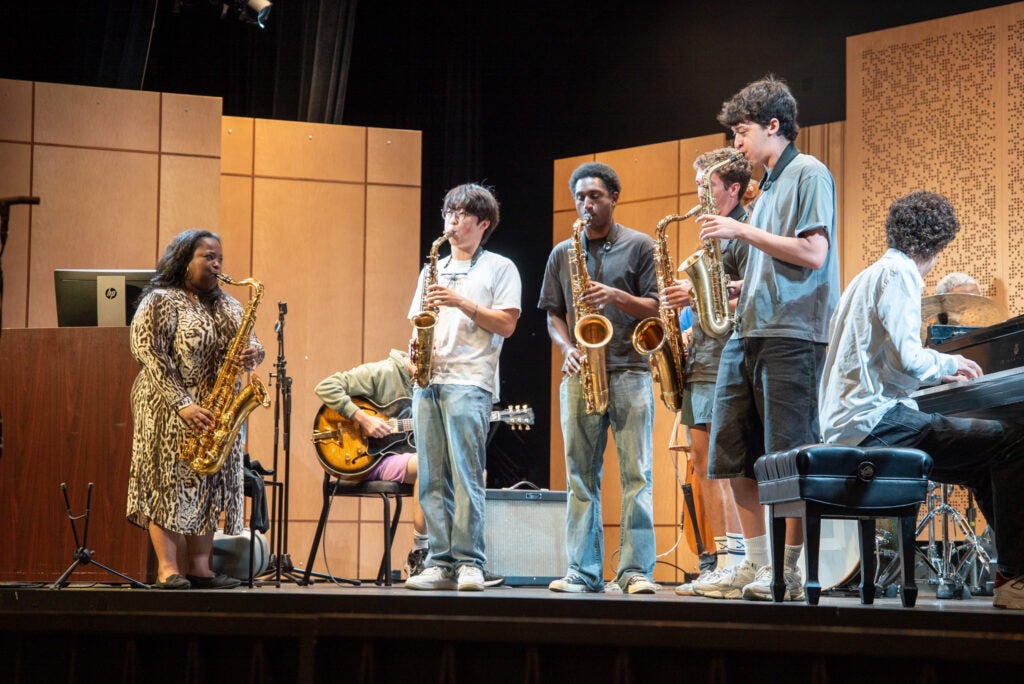 Three students with saxophones and one on piano on stage with a teacher instructing on saxophone.